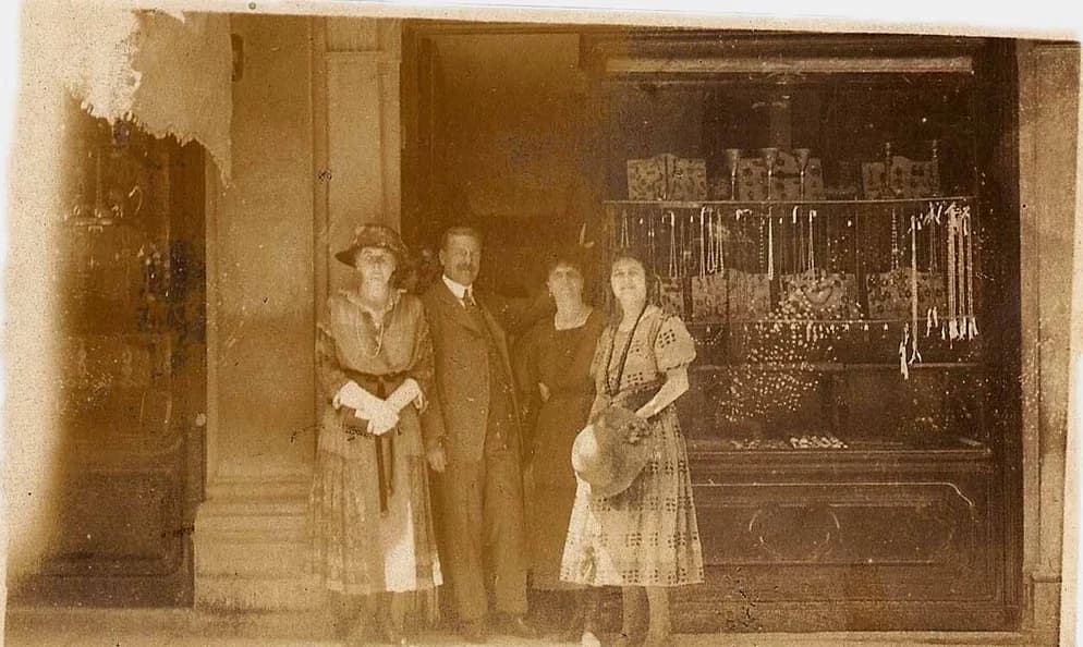 A sepia period photograph: a man and three women in 1920s dress in front of a jeweller’s window hung with necklaces